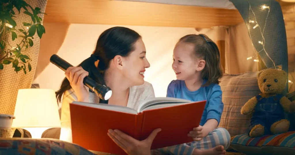 Effective Strategies To Improve Your kid's Reading Comprehension Skills 5 A mother and her young daughter reading a book with a flashlight in a cozy indoor tent setup, surrounded by soft lights, creating a magical and intimate reading atmosphere.