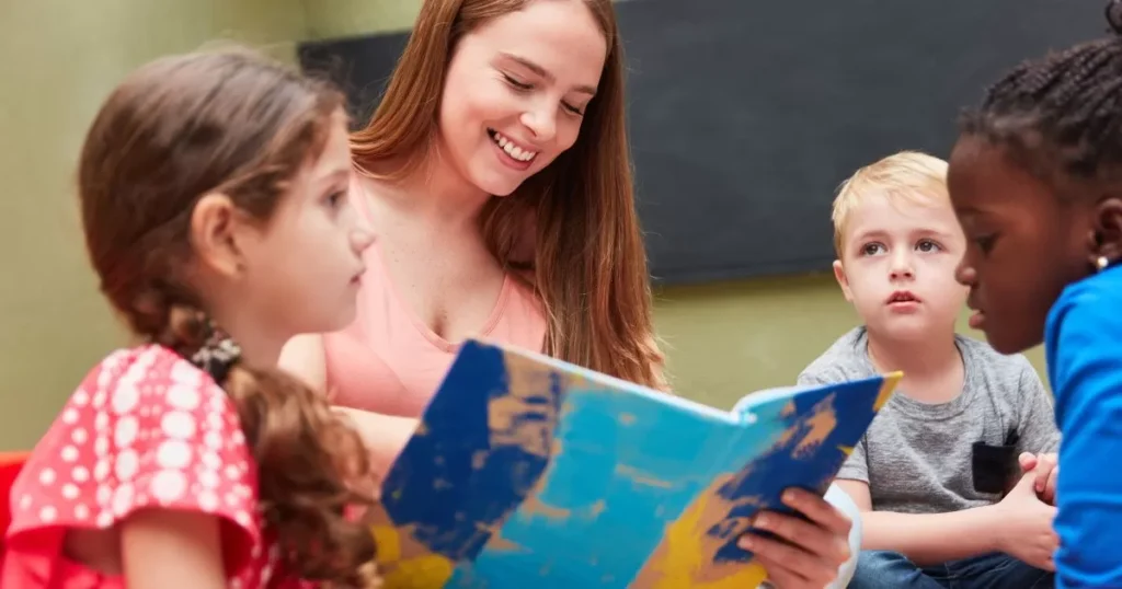 Effective Strategies To Improve Your kid's Reading Comprehension Skills 7 A teacher is reading a colorful book to a group of young children in a classroom. The children are gathered around, listening attentively, with expressions of curiosity and engagement. The scene reflects an interactive and supportive learning environment.