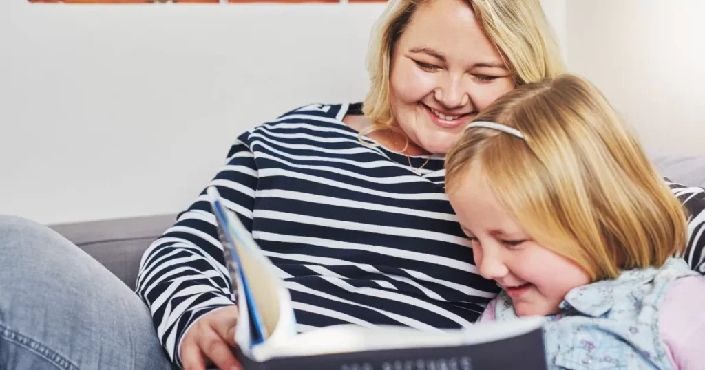 Effective Strategies To Improve Your kid's Reading Comprehension Skills 8 A mother and her young daughter are sitting together on a couch, smiling and enjoying a book. The mother looks on warmly while the daughter laughs, creating a joyful and cozy reading moment that highlights their bond and shared enjoyment of reading.